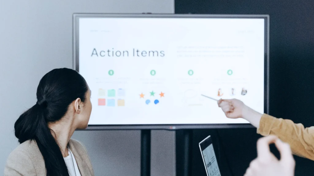 Das agile Framework Scrum: Auf einem Bildschirm werden verschiedene "Action Items" gezeigt. rechts zeigt eine Hand mit einem Stift auf den Bildschirm. Links vorne schaut eine Frau auf den Bildschirm im Hintergrund.
