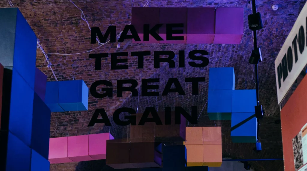 Kanban kann man sich vorstellen wie das Spiel Tetris: verschiedene bunte Tetris-Blöcke hängen in einer Halle an einer Decke aus roten Ziegelsteinen. Im Vordergrund steht auf einer Glasscheibe in schwarz: "Make Tetris great again".