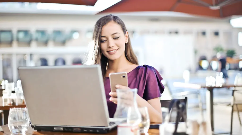 Termine in WooCommerce einfach verwalten: Eine Frau sitzt an einem Tisch vor ihrem Laptop. Sie hält ein Smartphone in der Hand und schaut darauf. Sie lächelt.