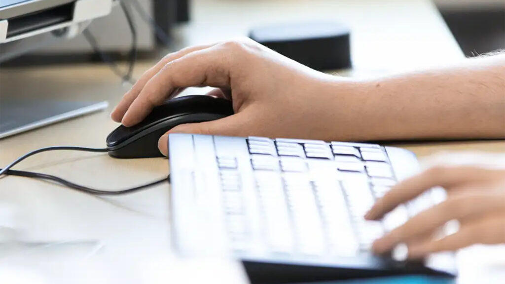 Man sieht die Hände von einer Person von der Seite mit der rechten Hand an einer Computermaus und der linken Hand an der Tastatur.