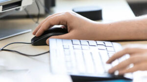Man sieht die Hände von einer Person von der Seite mit der rechten Hand an einer Computermaus und der linken Hand an der Tastatur.