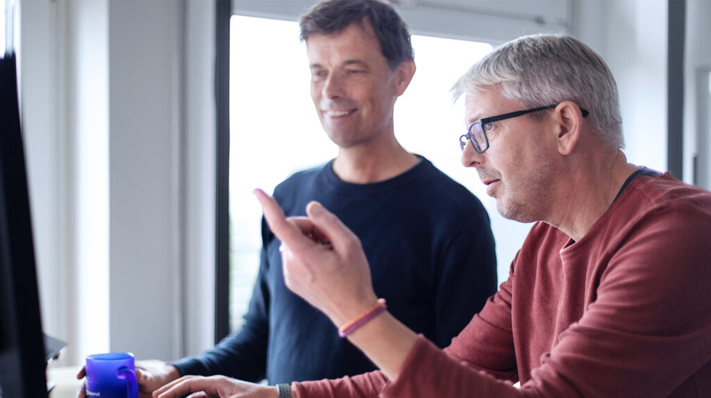 Zwei Männer mit einem grünen und rotem Pullover. Der Mann mit dem roten Pullover und der Brille zeigt mit dem Finger auf einen Desktop. Der Mann mit dem blauen Pullover hat eine blaue Tasse in der Hand und schaut auf den Desktop.