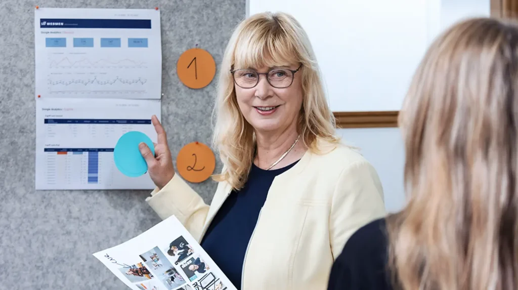 Auf dem Bild sieht man Christiane von Webmen. Sie zeigt mit dem Finger auf eine Tafel mit Statistiken drauf und schaut dabei Alena von Webmen an, die mit dem Rücken zur Kamera sitzt.